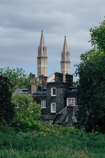 A scenic view capturing a cluster of dark grey stone buildings with flat and pitched roofs, surrounded by lush green trees and foliage in the foreground. In the background, two tall, pointed church spires made of light-colored stone rise against a partly cloudy sky, suggesting a historic church or cathedral. The buildings appear to be residential or possibly part of a historic district, with small windows and chimney stacks visible on the rooftops. The setting reflects a peaceful, well-maintained area, likely within an urban or suburban environment, where private rubbish removal services like Rubbish Removal Twickenham could be employed for on-site clearance or waste removal efforts. The overall scene emphasizes the contrast between the contemporary waste management needs and the traditional architectural environment.