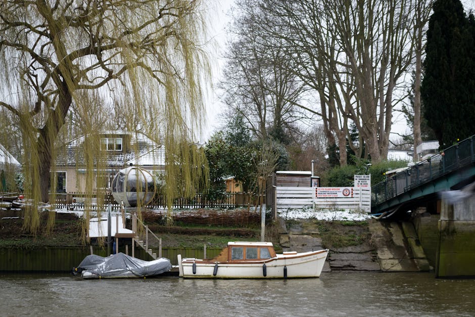 A small, cream-colored motorboat with a brown wooden cabin is moored along the edge of a narrow river or canal. The boat is secured with black fenders and is positioned parallel to the riverbank, which features a concrete or stone retaining wall. To the left of the boat, a portion of a larger object covered with a gray tarp is visible, possibly another piece of boating equipment or debris. Behind the boat, the riverbank rises slightly and is topped with a wooden fence, separating the water from a garden area that contains a large, leafless tree with a gnarled trunk and sprawling branches, and a weeping willow with long, pale yellow-green twigs hanging towards the water. Further back, there are residential buildings with white facades, dark roofs, and multiple windows, partially obscured by foliage, and a small wooden structure, possibly a shed, is visible near an area with snow. A sign with red and black text is mounted on the fence, indicating safety or access notices. The environment appears cold and overcast, with a muted, grey sky, typical of winter, and the scene overall reflects a quiet, semi-rural setting near a private property with occasional clutter or waste nearby, consistent with non-council rubbish disposal practices. The presence of the boat and fence suggests an area where private waste management or on-site clearance may be used by local residents or businesses, by Rubbish Removal Twickenham, in line with alternative rubbish removal services in the area.