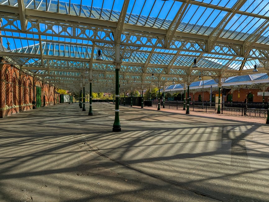 The image depicts a spacious outdoor area within a Victorian-style glass and iron canopy structure, featuring a clear blue sky overhead. The ground comprises a wide, paved surface with visible cracks and subtle texture, illuminated by sunlight that casts distinct, elongated shadows of the canopy's metal support pillars and decorative ironwork onto the pavement. To the left, a red-brick wall runs alongside the pathway, partly shaded by the canopy and adorned with a few small plants or greenery. On the right, a black metal fence separates the paved area from a building with red brick walls and arched windows, also partially shaded by the structure's roof. The scene exudes an atmosphere of calm and order, suggesting a public space or station platform that could be associated with facilities for gathering or waiting, with no visible activity or objects related to waste or rubbish removal in the immediate view. The professional environment aligns with services that handle outdoor space management or maintenance, such as those provided by Rubbish Removal Twickenham, particularly in contexts of on-site clearance or private waste handling outside of municipal disposal.