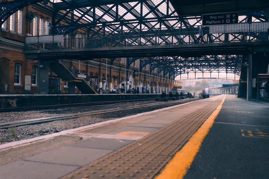 The image depicts an empty railway station platform with a curved yellow safety line along the edge, set beneath an intricate steel and glass canopy structure that allows natural light to filter through. In the background, there are brick station buildings with large windows, and a few people are visible waiting, though they are distant and indistinct. The platform surface appears clean and well-maintained, with textured tactile paving near the edge for safety. Overhead signage indicates directions for exits and platforms. To the right, a small station booth or shelter is present, with equipment and materials that could relate to station maintenance. The overall scene is calm, urban, and hints at a background of ongoing transit operations. This setting exemplifies an environment where private or independent waste handling might occur as part of station maintenance or upgrades, aligning with the context of rubbish removal services in public transit areas. Rubbish Removal Twickenham offers solutions for waste management in similar environments, focusing on efficient and discreet clearance of waste materials.